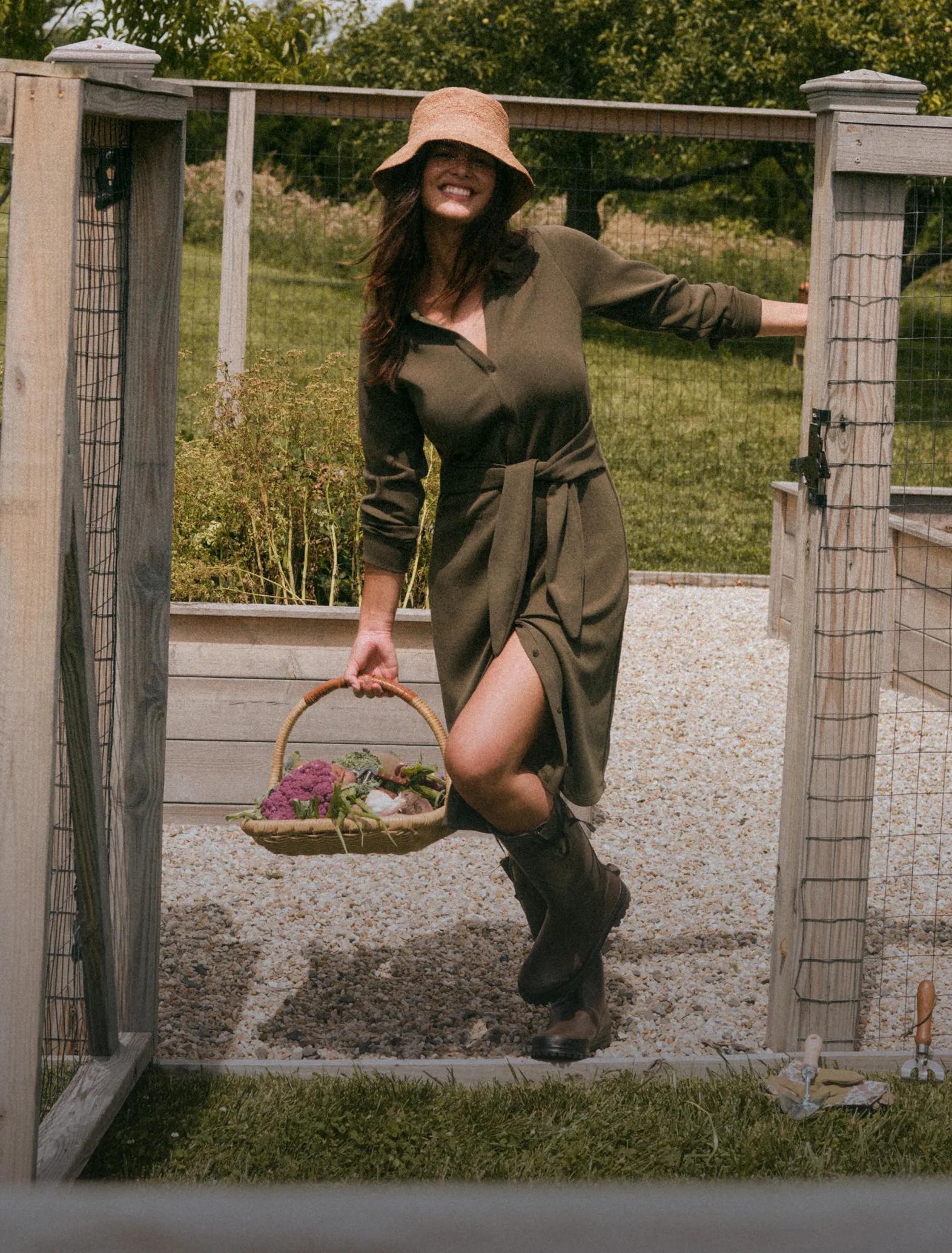 Model in an olive green long-sleeve dress with a side slit and a wide-brim tan hat carries a floral basket while walking through a garden gate.