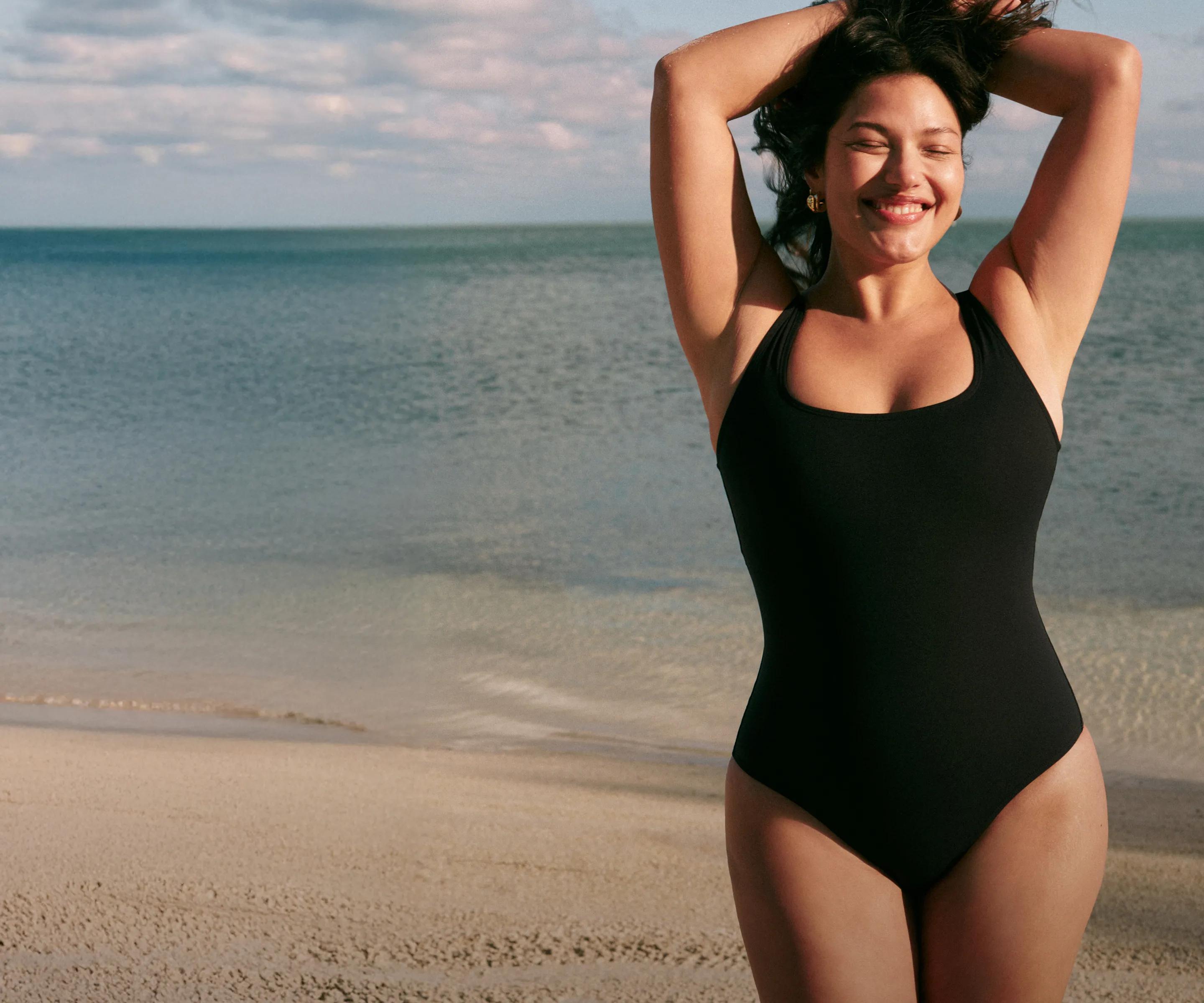 Model wearing a black scoop-neck one-piece swimsuit with a racerback, standing on a sandy beach with the ocean behind her