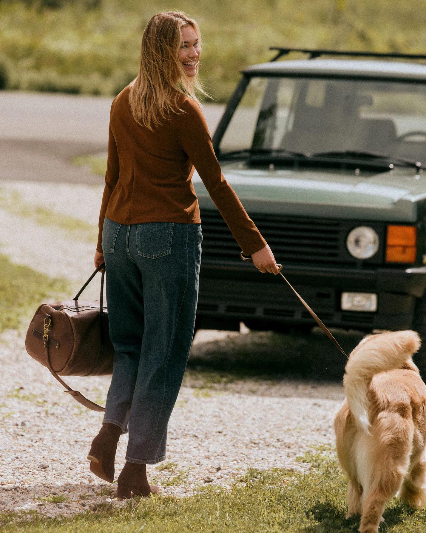 Model in a rust-colored sweater and wide-leg jeans walks a golden retriever on a leash while carrying a brown duffel bag, with a green SUV in the background.
