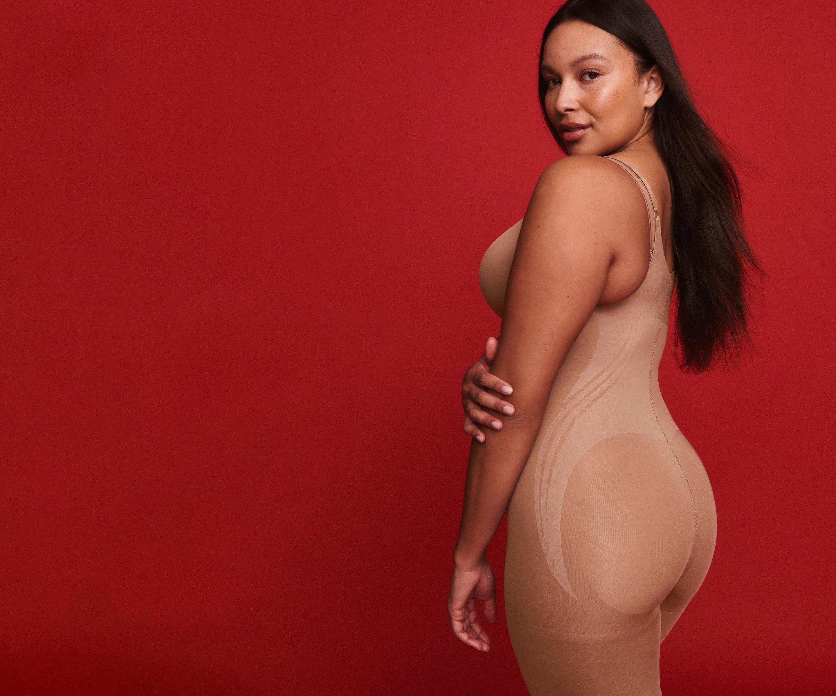 Model against a red background wears a nude thin-strap shapewear bodysuit, shown from the side highlighting the smoothing silhouette.