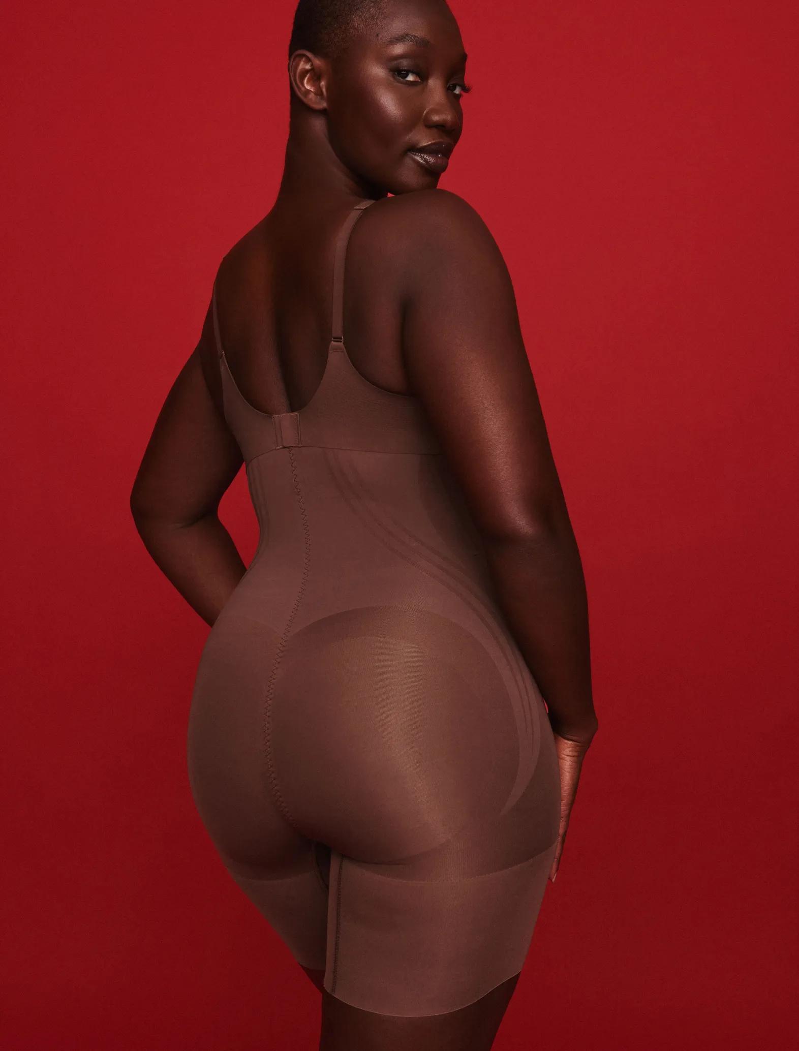 Model wearing a cocoa brown strapless bodysuit shaper with mid-thigh coverage against a bold red background