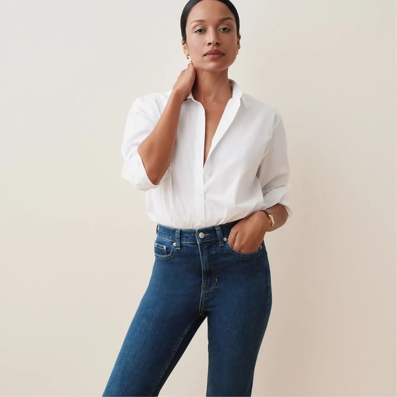 Model touches her neck, wearing a white button-down collared shirt with rolled sleeves and medium-wash high-rise jeans.