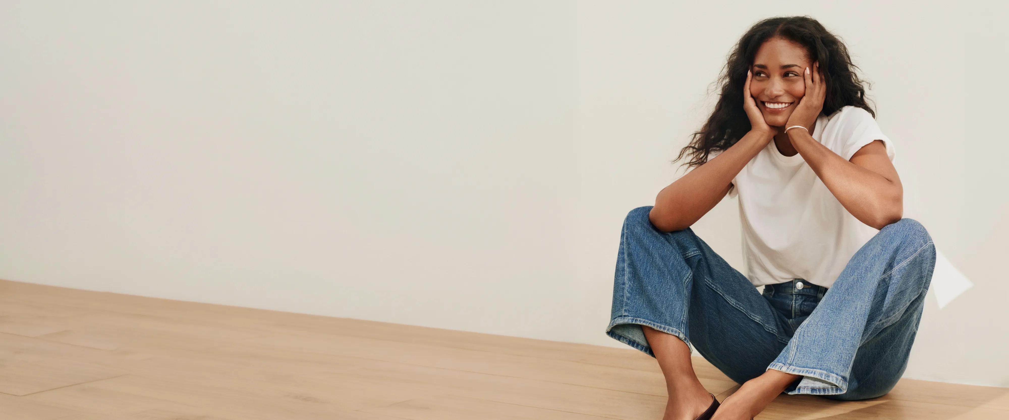 Model sits smiling on a wooden floor, resting her chin on her hands, wearing a white t-shirt and medium-wash wide-leg jeans.