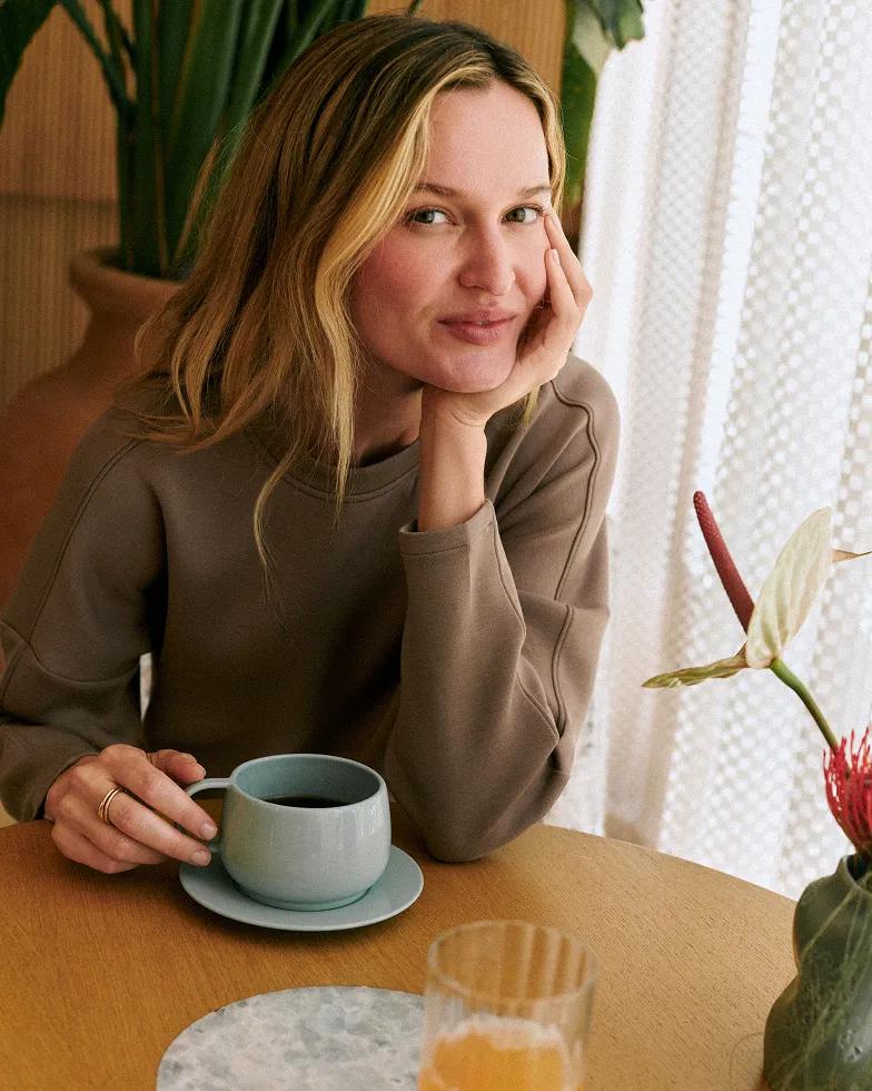 Woman wearing a taupe cozy sweatshirt smiling at a wooden table with a cup of coffee, seated near a vase of flowers by a sunlit window.