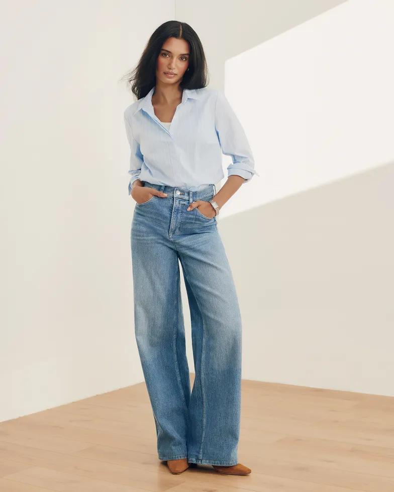 Model wears a light blue collared button-down shirt with rolled sleeves, paired with light-wash high-rise wide-leg jeans and tan pointed-toe heels.