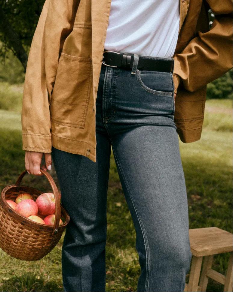 Vue partielle d'une personne portant une veste beige sur un t-shirt blanc et un jean bleu foncé avec une ceinture noire, et transportant un panier tressé rempli de pommes en extérieur.