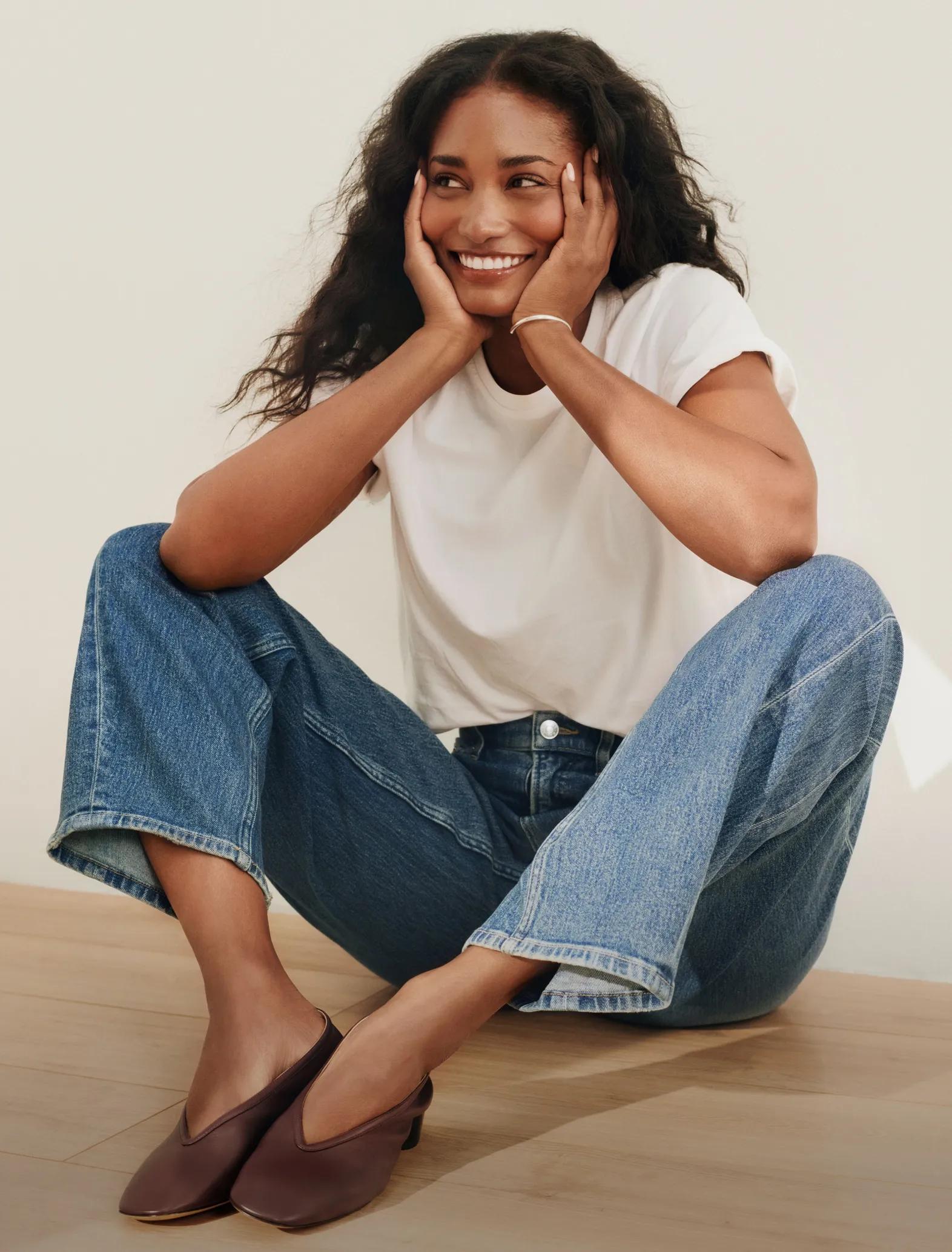 Model sits smiling on a wooden floor, resting her chin on her hands, wearing a white t-shirt and medium-wash wide-leg jeans.
