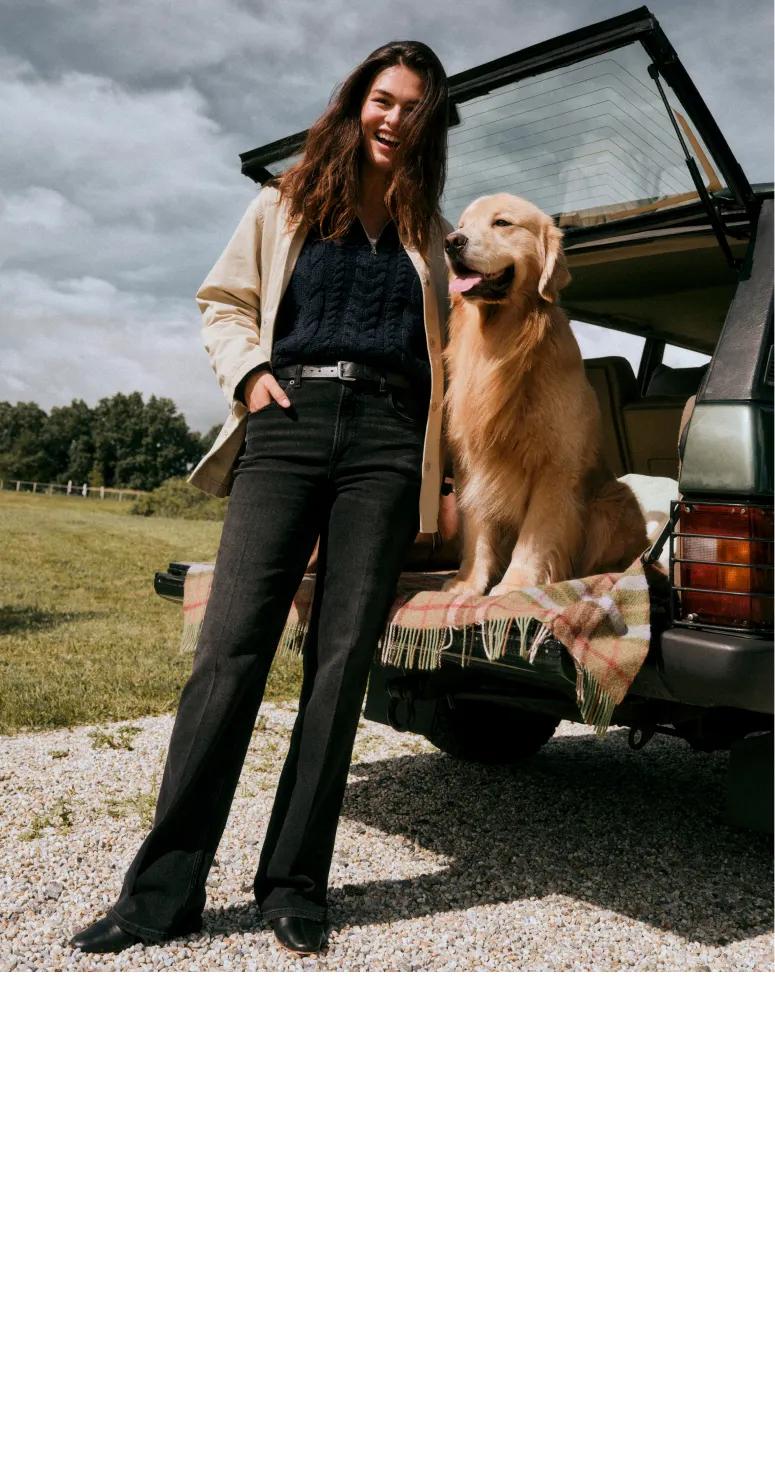Model stands beside a golden retriever seated in the back of an SUV, wearing a cream jacket, navy sweater, and black flared jeans.