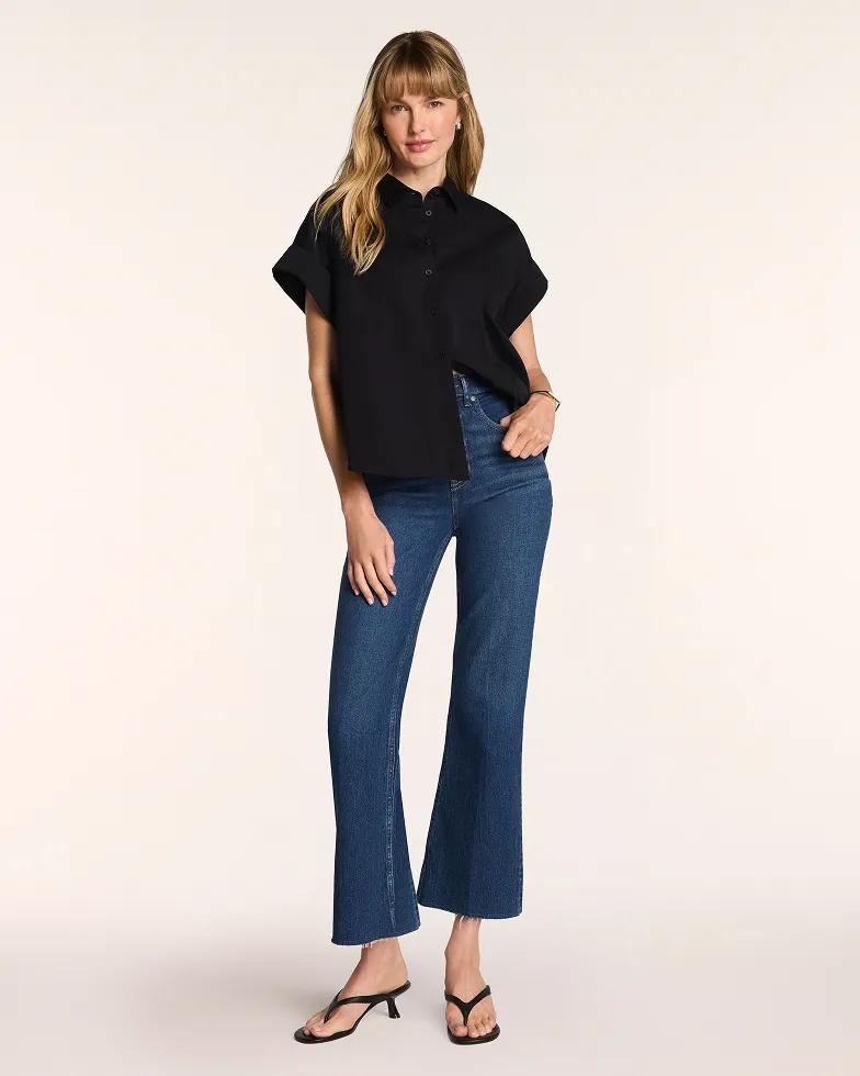 Model wears a black collared short-sleeve top with dolman sleeves, paired with medium-wash flared jeans and black strappy heeled sandals.