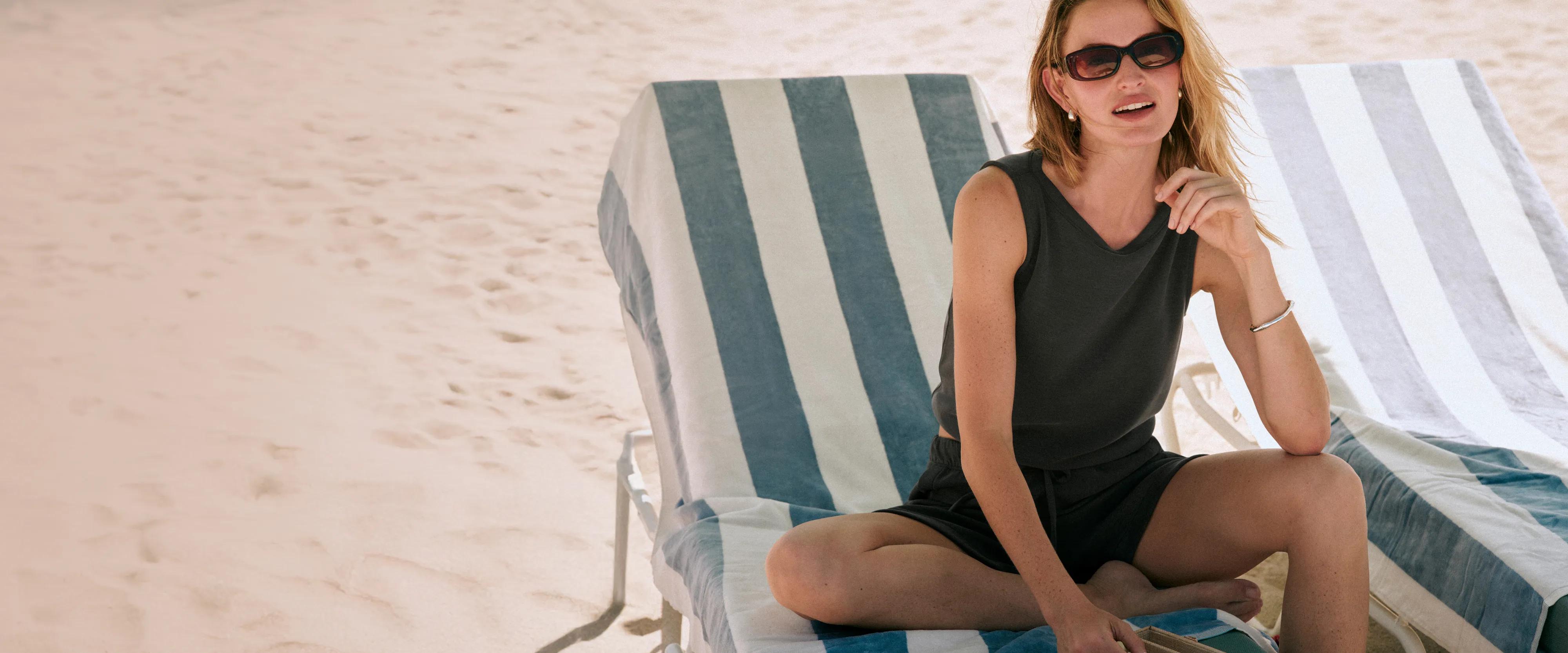Model wearing a dark olive sleeveless romper with button detail, seated in a beach chair on the sand
