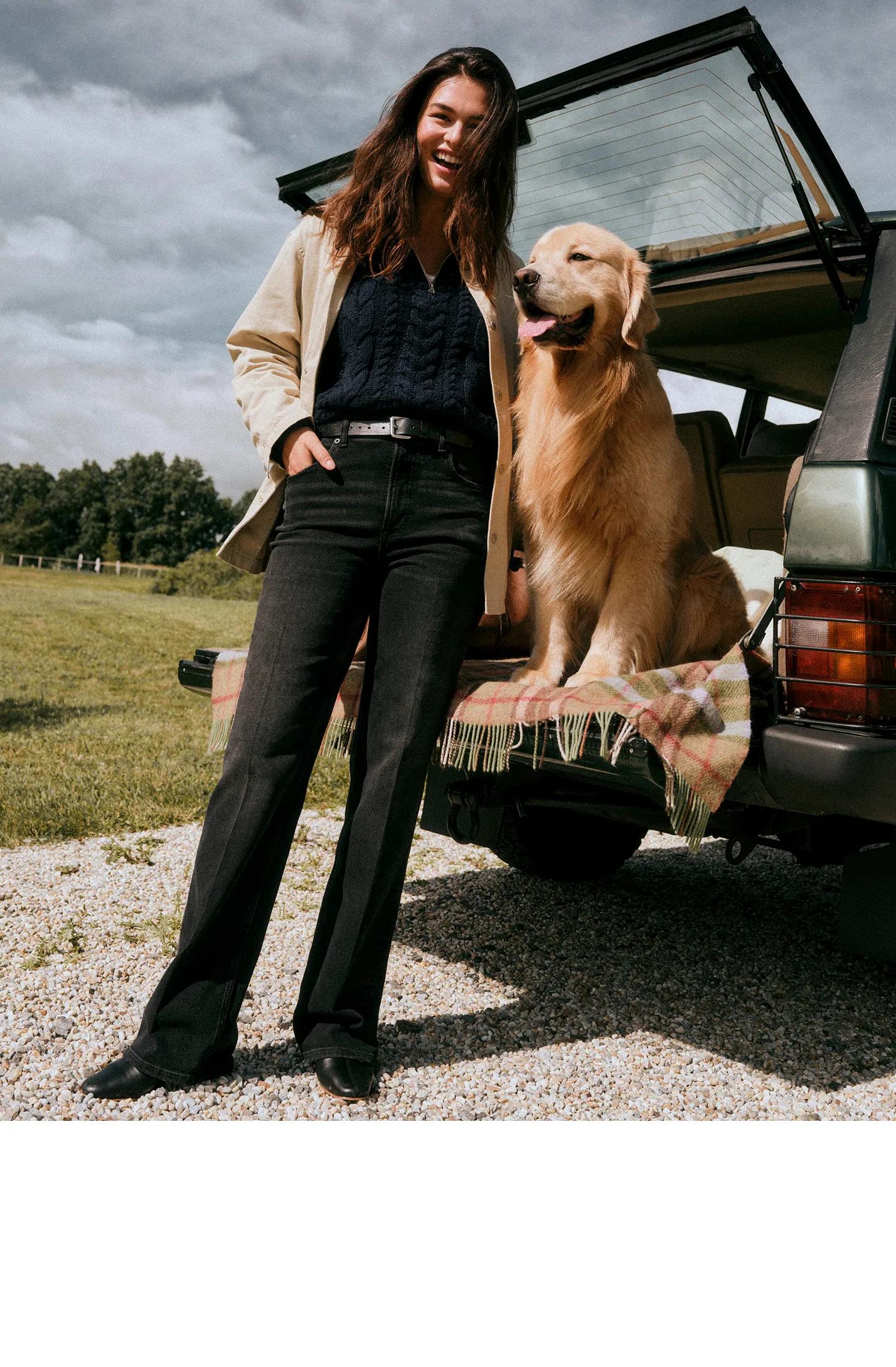 Model stands beside a golden retriever seated in the back of an SUV, wearing a cream jacket, navy sweater, and black flared jeans.