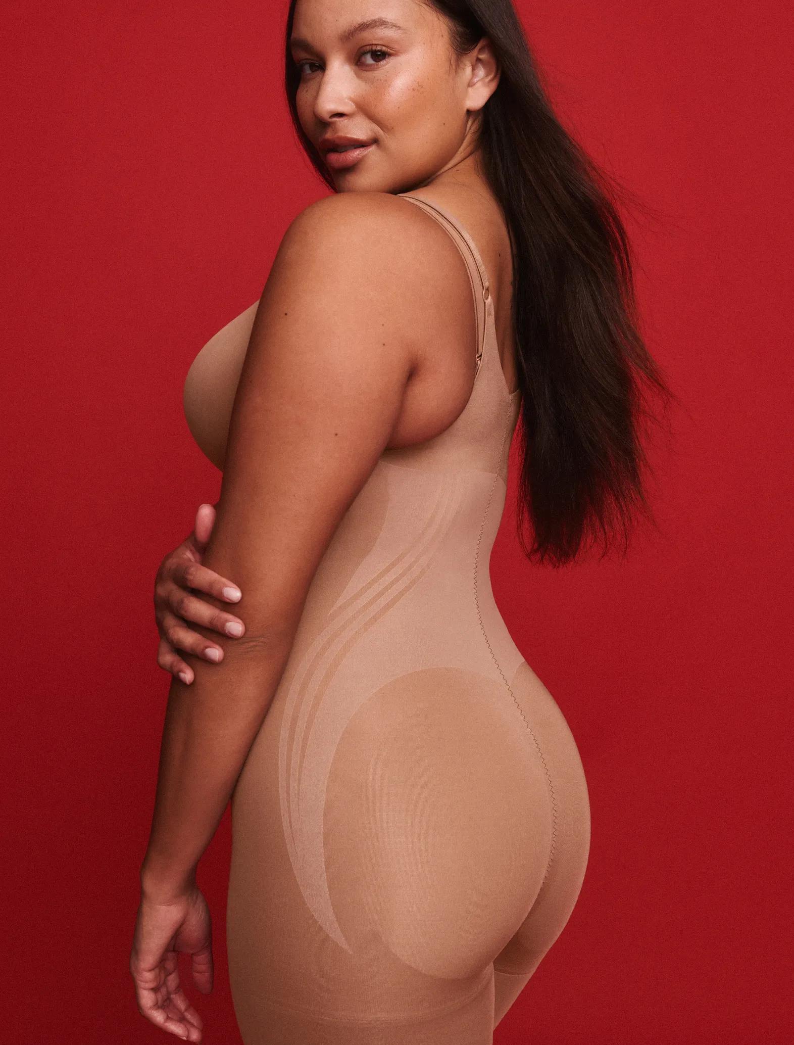 Model against a red background wears a nude thin-strap shapewear bodysuit, shown from the side highlighting the smoothing silhouette.