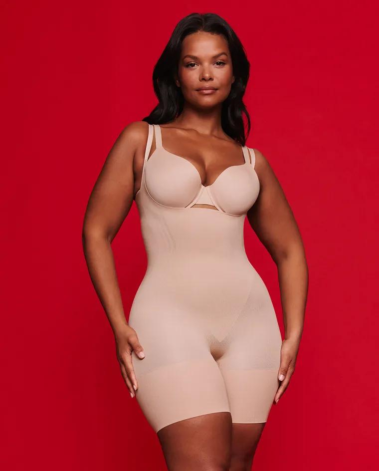 Model wearing a nude open-bust shaping bodysuit with mid-thigh coverage and double shoulder straps against a red background