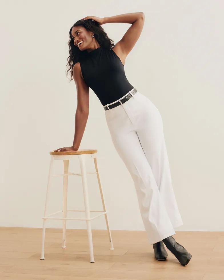 Model laughs while leaning on a stool, wearing a black sleeveless mock-neck top with white belted trousers and black pointed-toe boots.