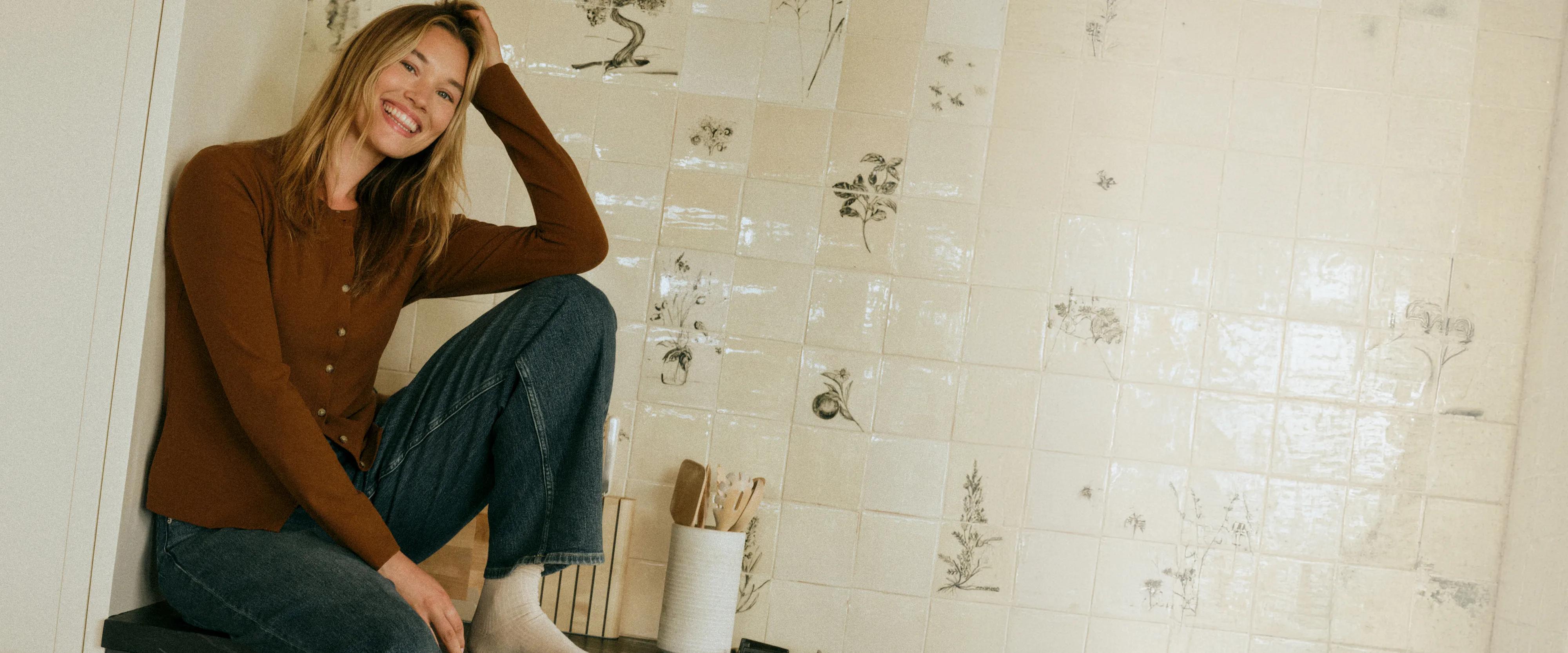 Model smiles while sitting against a decorative tiled wall, wearing a rust cardigan and medium-wash jeans.