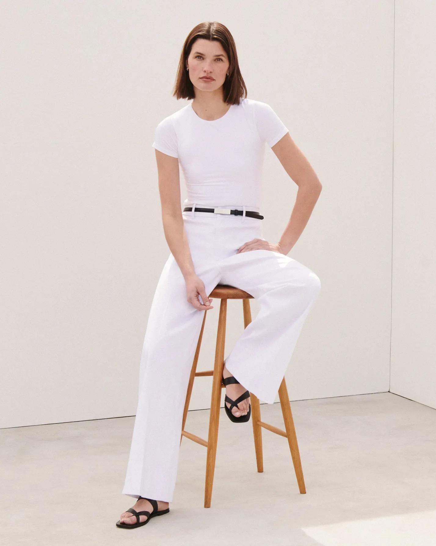 Model seated on a stool wearing white wide-leg pants and a fitted white crewneck tee, styled with a slim black belt and black strappy sandals