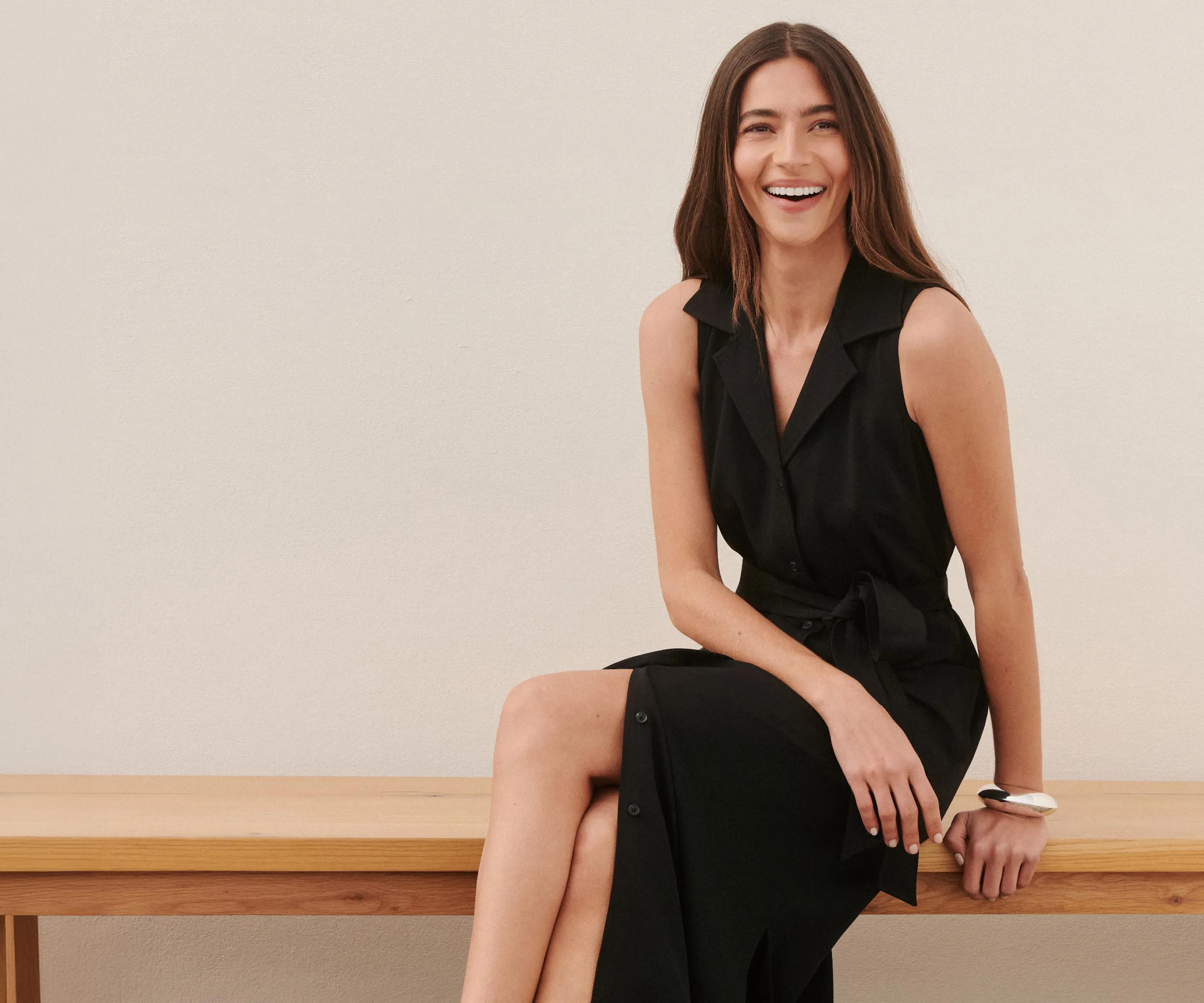 Model seated on a bench wearing a black sleeveless midi dress with a notch collar, tie waist, button-front placket, and side slit