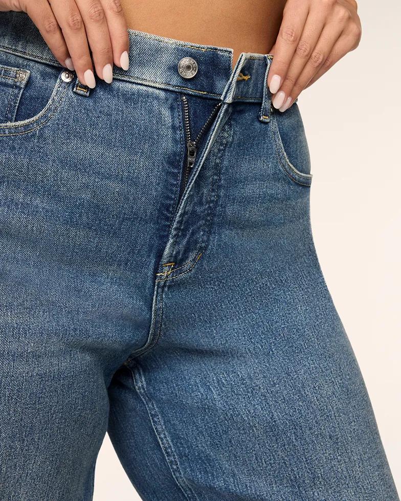 Close-up of a model's hands unfastening the button-fly closure on a pair of medium-wash denim jeans.
