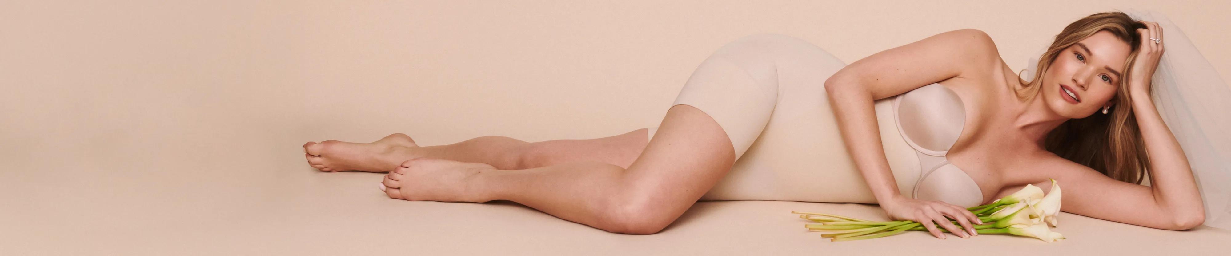 Model wearing a nude strapless bra and mid-thigh shaping shorts, reclining with white calla lilies against a warm beige backdrop