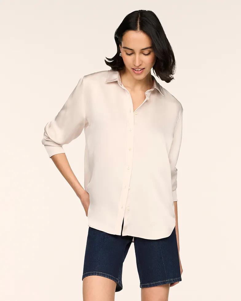 Model wears a cream satin button-down shirt with a collared neckline and rolled sleeves, paired with dark denim shorts.