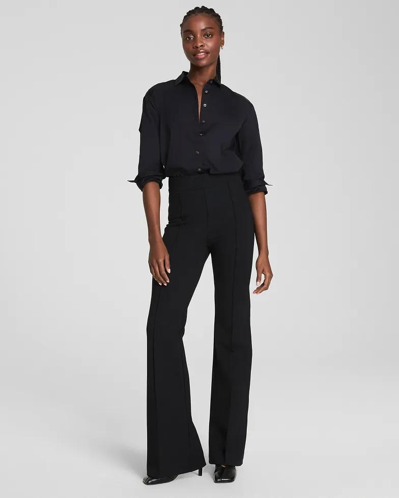 Model wears an all-black satin button-down shirt with rolled sleeves and matching high-waisted flared trousers with black pointed-toe heels.