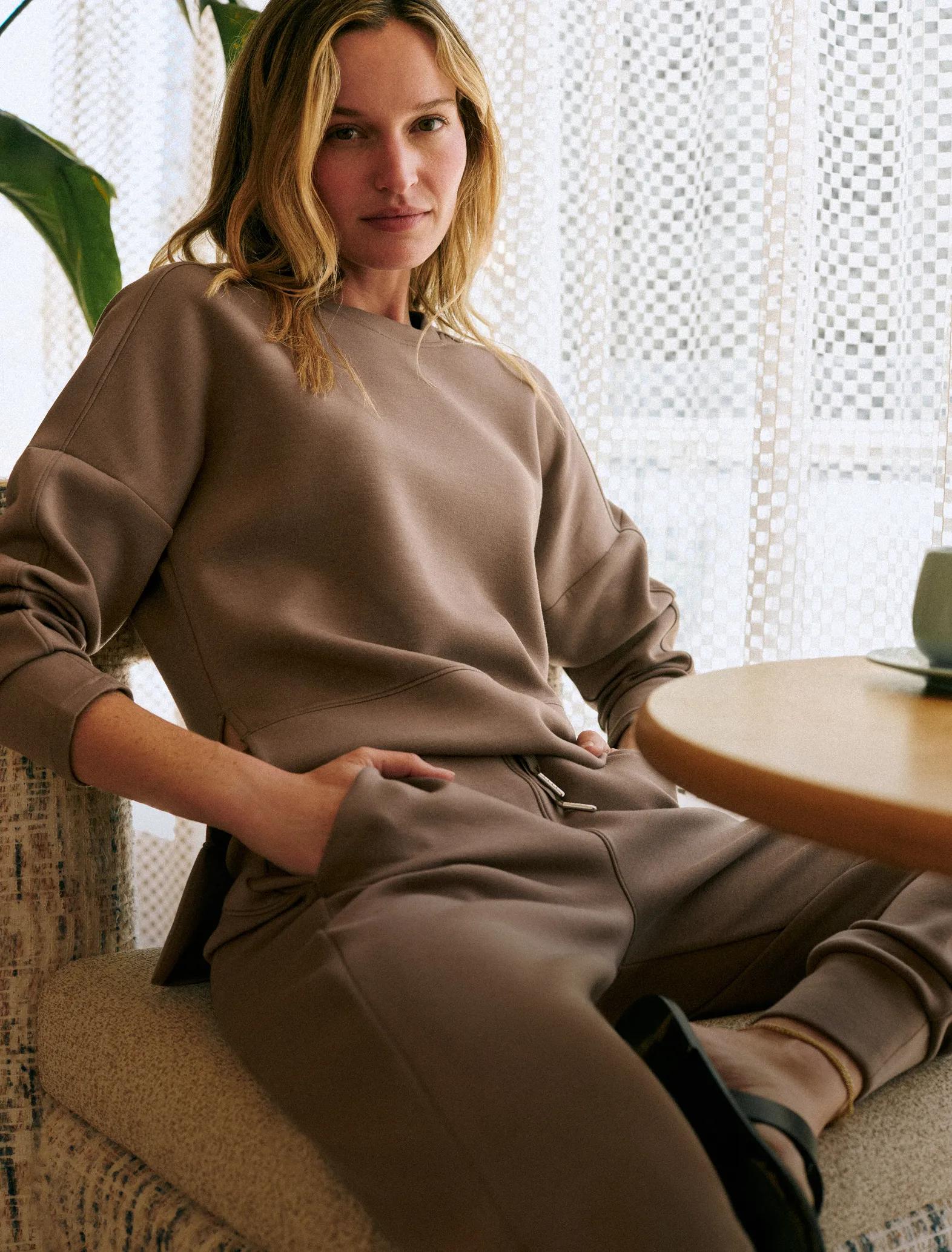 Woman seated on a boucle chair wearing a taupe cozy sweatshirt and jogger set, next to a wooden side table by a sunlit window.