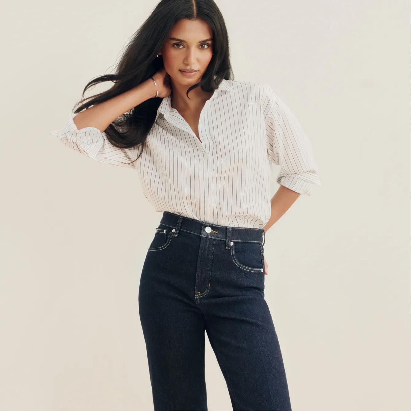 Model runs a hand through her hair, wearing a white pinstripe button-down shirt paired with dark-wash high-rise jeans.