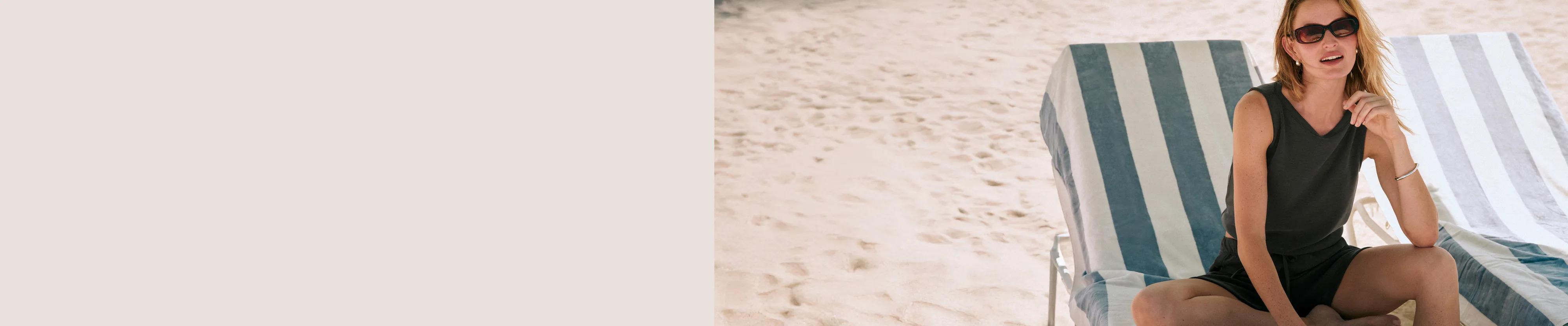 Model wearing a dark olive sleeveless romper with button detail, seated in a beach chair on the sand