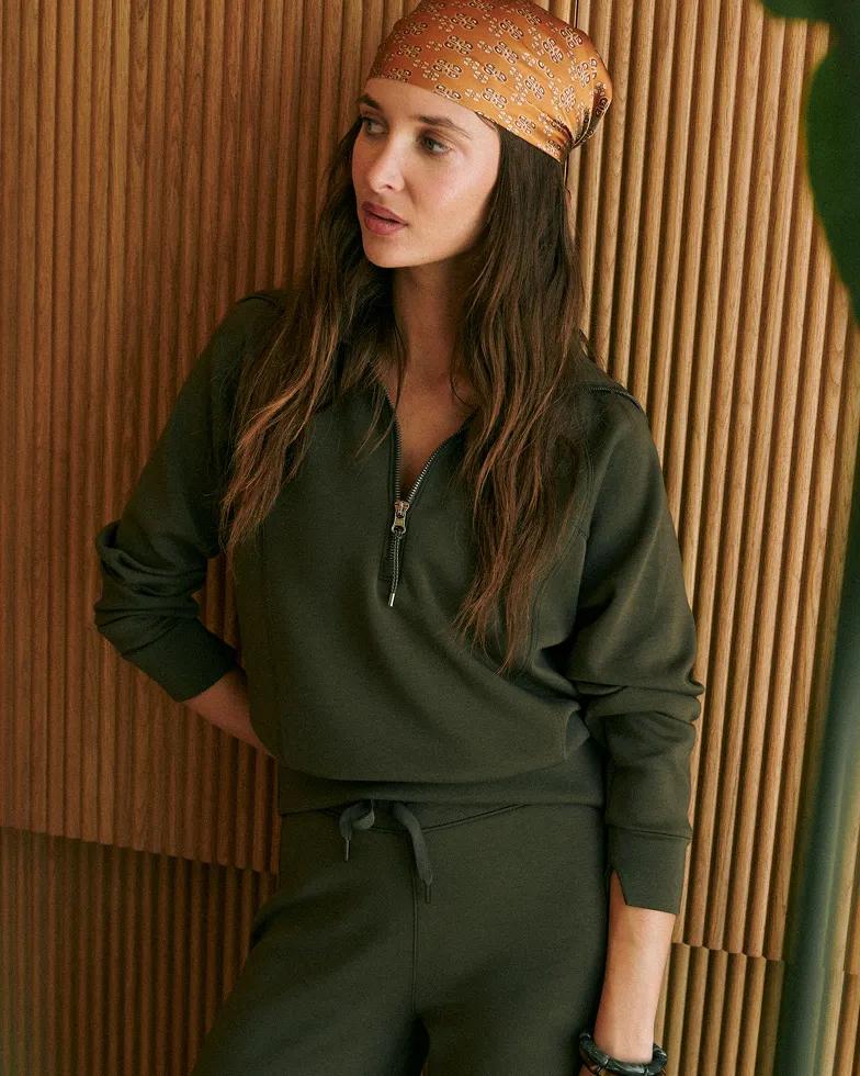 Model wears a patterned orange headscarf and an olive green zip-front collared sweatshirt with matching drawstring pants, leaning against a wood-paneled wall.