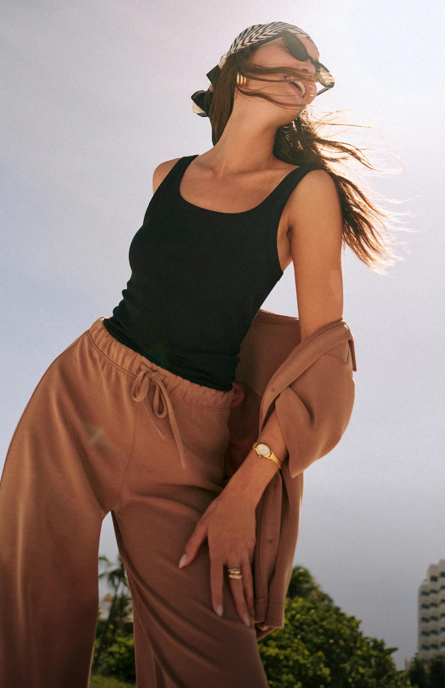 Model wearing a black scoop-neck tank and terracotta drawstring wide-leg pants, with a matching jacket draped over one arm
