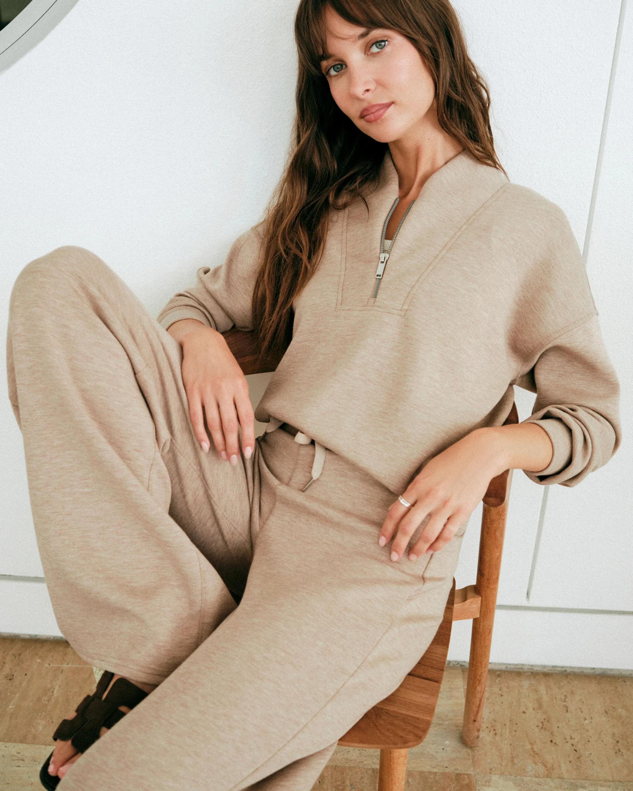 Model wearing a oatmeal heathered quarter-zip pullover and matching jogger pants, seated on a wooden chair