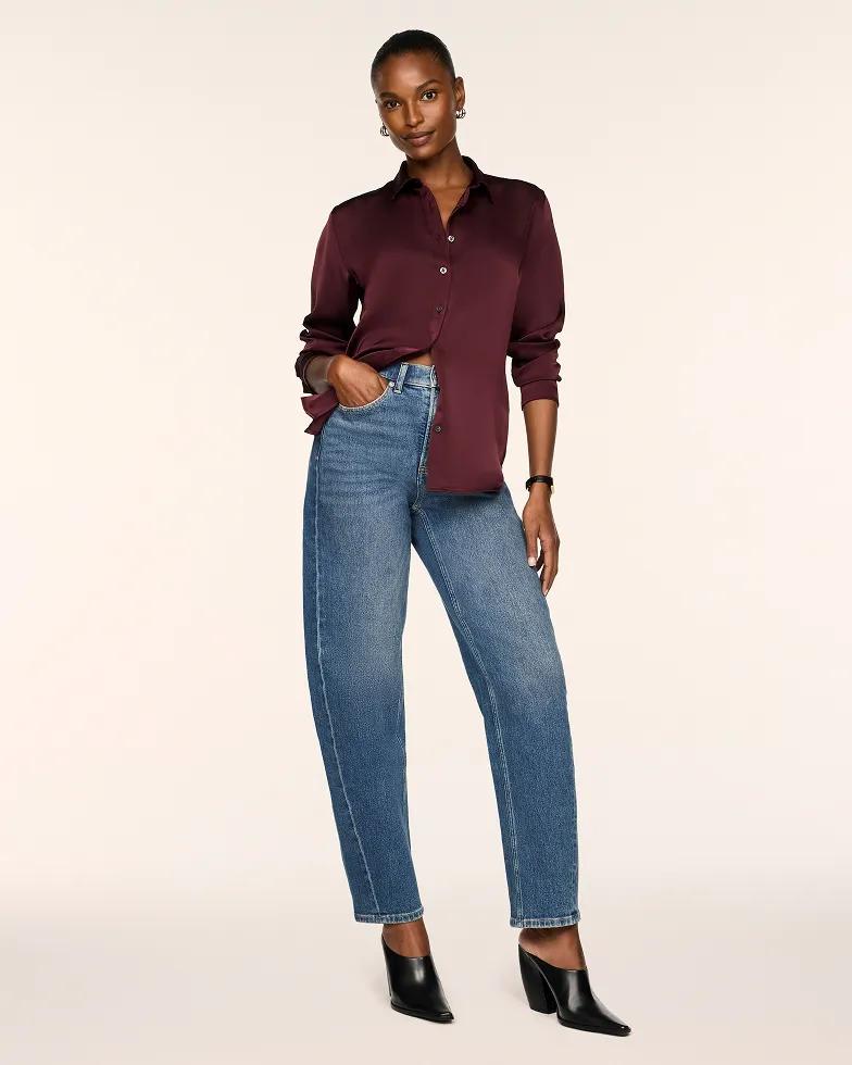 Model wears a burgundy satin button-down blouse with a collared neckline, paired with medium-wash straight-leg jeans and black pointed-toe heeled booties.