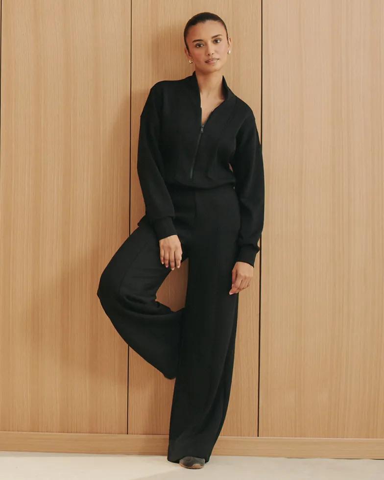 Model leans against a wood-paneled wall wearing a black zip-front mock-neck top with ribbed cuffs and matching wide-leg pants.