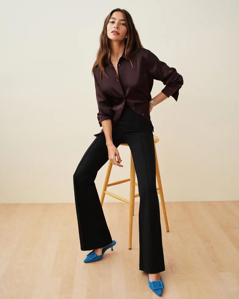 Model leans against a yellow stool wearing a dark brown satin button-down shirt with black flared trousers and blue pointed-toe heels.