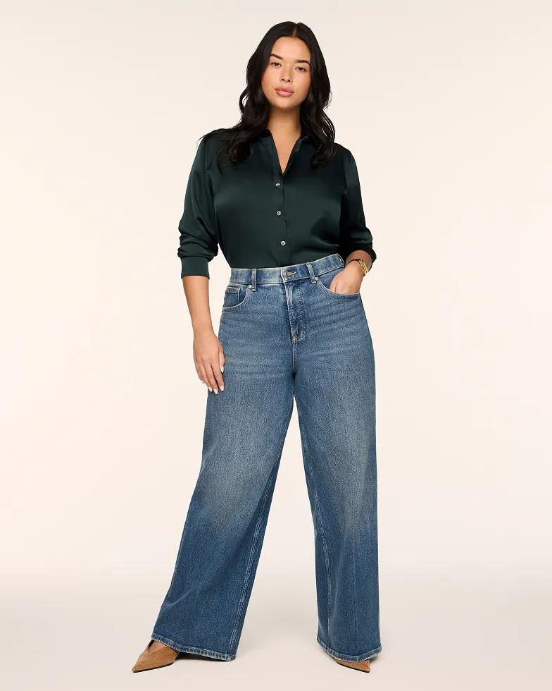 Model wears a dark green satin button-down blouse with a collared neckline, paired with medium-wash high-rise wide-leg jeans and tan pointed-toe heels.