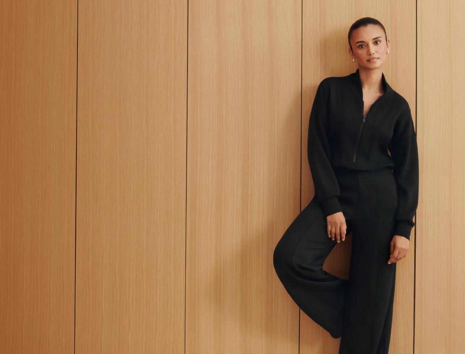 Model leans against a wood-paneled wall wearing an all-black long-sleeve jumpsuit.
